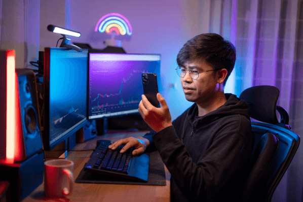 Mann vor Computer kontrolliert Kurs in Kryptobörse auf Smartphone