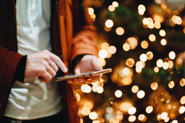 Haende, die ein Handy mit Weihnachtskampagnen darauf halten, mit einem Weihnachtsbaum im Hintergrund