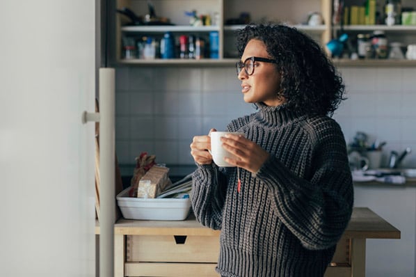 An Winterdepression leidende Frau steht in Küche mit Teetasse in Händen