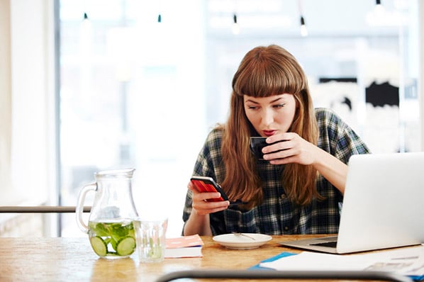 Frau als Workaholic sitzt am Laptop, trinkt und schaut auf Smartphone