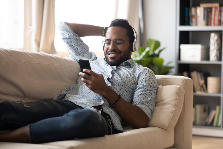 Mann mit Kopfhörern liegt auf der Couch und hält Smartphone in der Hand