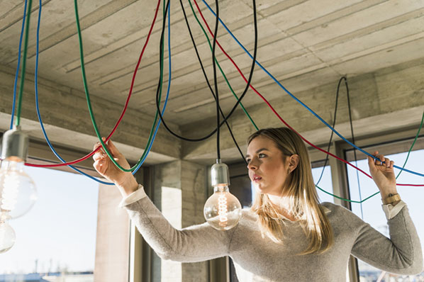 Junge Frau schaut auf Kabel an Decke vor Glühbirne symbolisiert Energiesparen im Büro