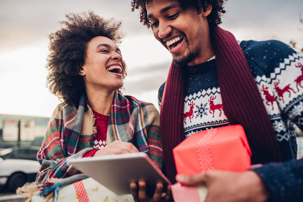 Lachende Frau und Mann in Weihnachtskleidung shoppen auf Tablet online im Weihnachtsgeschäft