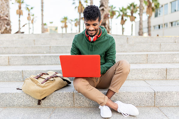 Junger Mann auf Steintreppe draußen synchronisiert Google-Kalender mit Outlook auf rotem Laptop